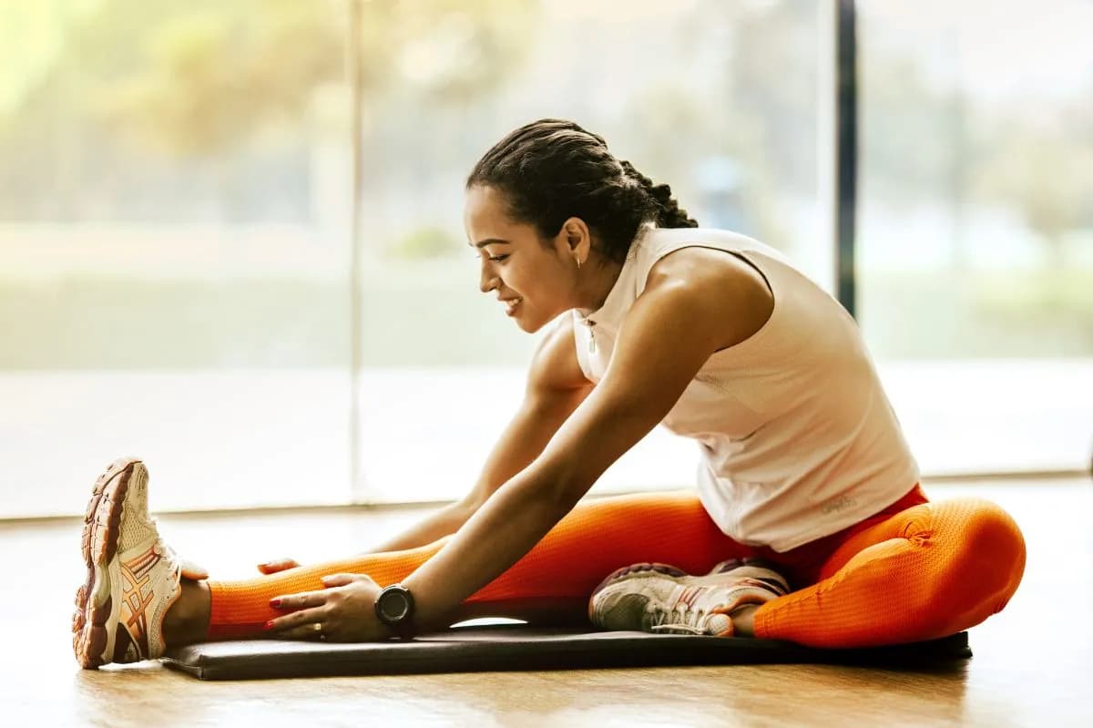 Woman doing a seated forward stretch on a mat by a bright window